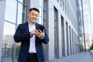 A smiling young Asian male businessman in a suit is standing outside an office center and using a mobile phone. photo