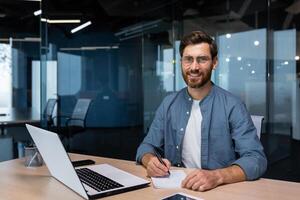 retrato de exitoso empresario detrás papel trabajar, hombre en camisa sonriente y mirando a cámara, financiero jefe dentro oficina utilizando ordenador portátil en trabajo a lugar de trabajo. foto