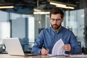 A young serious business man is sitting in the office at a desk with a laptop and is focused on working with documents. Writes and checks data and invoices. photo