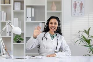 un alegre hembra médico atractivo en un llamar, ondulación a el cámara desde su oficina, equipado con médico literatura y plantas. foto