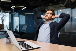 Completion work important project, mature man working inside modern office building using laptop, senior boss with beard and glasses putting hands behind head dreaming and visualizing future plans. photo