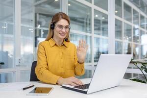 Office worker works remotely, online customer support service, help service, woman uses headset and laptop for online consultation and call, works inside office, waves greeting gesture. photo