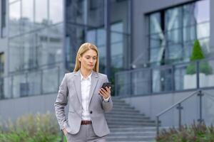 Young businesswoman in a suit standing near an office center and struggling with a mobile phone, writing a message, reading the news, chatting. photo