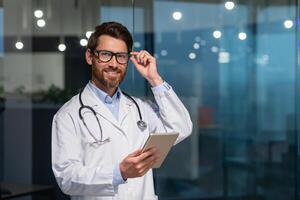 Portrait of a young handsome male doctor. He stands in the office in a white coat and with a stethoscope. He holds a tablet in his hands. He looks at the camera, adjusts his glasses, smiles. photo