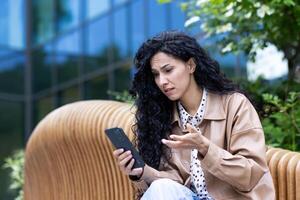 Thinking and upset woman outside office building with phone, Hispanic business woman frustrated reading news online and browsing internet pages, using smartphone. photo