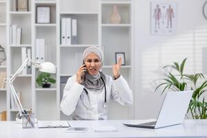 un hembra médico en un hijab aparece estresado mientras hablando en el teléfono y gesticulando con su otro mano en un bien equipado moderno oficina. foto