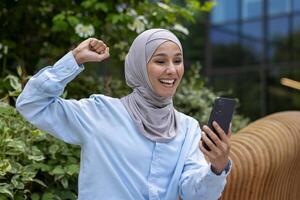 exitoso alegre musulmán mujer en hijab recibido en línea ganar notificación, dinka leyendo Noticias y participación mano arriba triunfo gesto celebrando sentado en banco fuera de oficina edificio en parque. foto