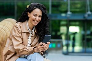 Professional young Hispanic businesswoman enjoying a break outside an office building, happily interacting with her smartphone. photo