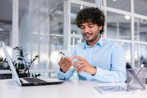 joven indio programador dentro moderno oficina sentado a lugar de trabajo, hombre utilizando teléfono, pruebas en línea solicitud en nuevo software, empresario empresario en camisa sonriente y contento con logro. foto