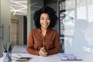 profesional joven mujer con Rizado cabello, en un punteado camisa, se involucra directamente con el cámara en un brillante, moderno oficina ajuste. foto