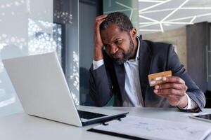 Upset and worried businessman at workplace, man cheated received online money transfer error, african american man unhappy sitting with laptop inside office holding bank credit card. photo