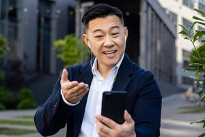 Close-up photo of a young Asian man in a suit and headphones, smiling and talking on the phone on a call.