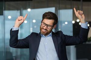 Mature successful manager boss celebrating good achievement and good work result, man dancing at workplace inside office, businessman at work in business suit. photo