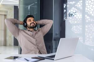 Close-up photo of a young Indian man sitting in an office at a desk and resting. Wearing headphones, he listens to music relaxed with his hands behind his head.