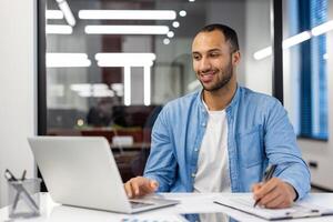 sonriente joven masculino Hispano estudiante sentado en moderno oficina a escritorio con ordenador portátil y haciendo notas en computadora portátil, distancia aprendiendo. foto