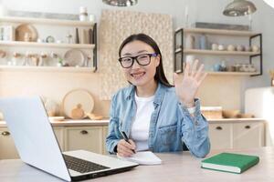 joven asiático hembra estudiante estudiando remotamente a hogar, retrato de mujer con ordenador portátil en en línea aprendizaje utilizando ordenador portátil mientras sentado en cocina a hogar foto