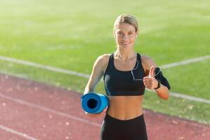 Smiling athletic woman holding a yoga mat and giving a thumbs up on a sunny track field, showcasing fitness and health. photo