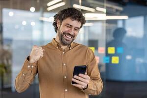 alegre masculino empresario emocionado zapatillas puño en victoria como él mira a su teléfono inteligente, en pie en un moderno oficina ajuste. foto