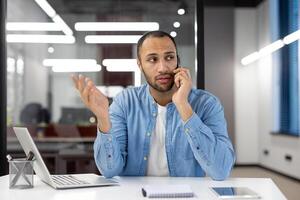 A man is talking on his cell phone while sitting at a desk. He is wearing a blue shirt and he is in a serious mood photo