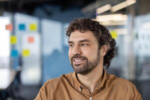 Portrait of startup creator looking aside with friendly smile on background of glassy walls reflecting light. Handsome entrepreneur listening to presentation of colleague about future innovations. photo