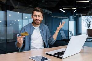 Portrait of disappointed and deceived businessman inside office, mature man looking at camera confused holding bank credit card and phone, employee got rejection error. photo