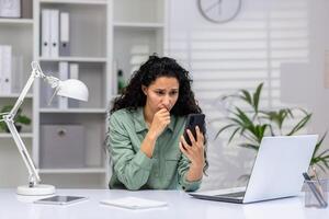 Upset young hispanic woman sitting in the office at the desk and looking worriedly at the phone screen. photo