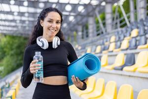 Fit, happy woman holding a yoga mat and a bottle of water, ready for a workout session in a stadium setting. photo