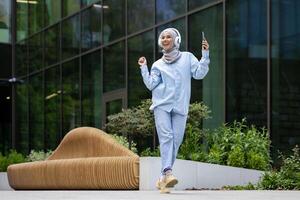 joven musulmán hembra estudiante fuera de Universidad instalaciones, contento bailando fuera de edificio, mujer en hijab escuchando a música utilizando teléfono y auriculares, contento sonriente. foto