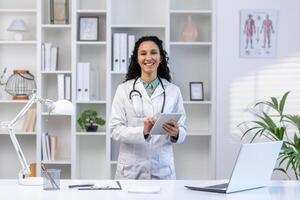 retrato de un hembra médico dentro el oficina de el clínica, el mujer es sonriente y mirando a el cámara con un tableta computadora en su manos, en pie a el escritorio, consultante pacientes remotamente foto