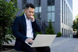 trastornado joven empresario sentado en un banco cerca un oficina centro, participación un ordenador portátil en su regazo y mirando preocupadamente a el pantalla. foto