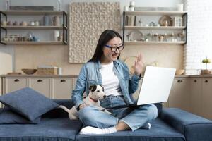 joven hermosa asiático mujer a hogar sentado en sofá y hablando en llamada utilizando computadora portátil, mujer con mascota perrito Jack Russell terrier hablando con amigos remotamente foto