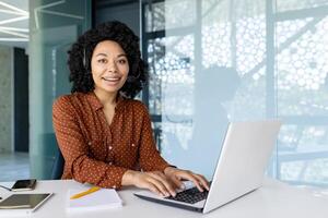 Portrait of female office worker with call headset and laptop inside office at workplace, african american smiling and looking at camera, woman satisfied with work and achievement results. photo