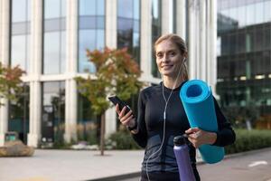 A woman gearing up for a fitness session holds her yoga mat and water in an urban setting while engaging with her smartphone, possibly curating a workout playlist or scheduling a wellness session. photo