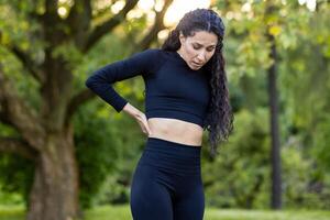 A Hispanic woman experiencing side pain while exercising in a green park. Her expression shows discomfort as she pauses her fitness routine. photo