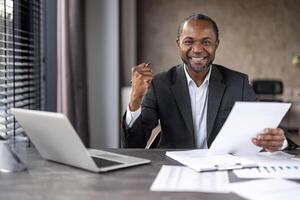 Successful African American businessman smiles while reviewing reports at his office desk with laptop and paperwork. photo