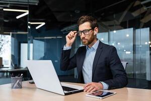 Mature serious thinking businessman working inside office, boss investor in business suit working at table with laptop brainstorming business strategy and plan. photo