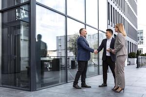 Meeting of three successful business people, diverse dream team man and woman outside office building, greeting and shaking hands, experienced professionals specialists in business suits talking photo