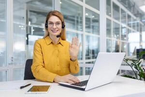 Office worker works remotely, online customer support service, help service, woman uses headset and laptop for online consultation and call, works inside office, waves greeting gesture. photo