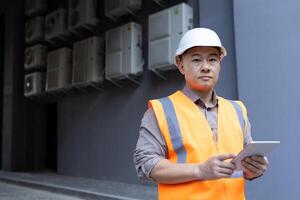 de cerca retrato de un joven asiático hombre vistiendo un difícil sombrero y chaleco, en pie fuera de un fábrica, construcción sitio, participación un tableta y mirando seriamente dentro el cámara. foto