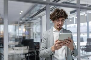 un grave y enfocado joven hombre es en pie en un oficina centrar y trabajando en un proyecto utilizando un tableta. foto