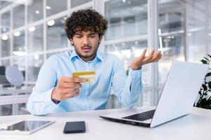problemas con el cuenta, pago, transacción. un joven musulmán empresario es sentado en el oficina a el ordenador portátil y mirando preocupadamente a el crédito tarjeta. foto
