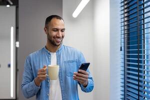 sonriente joven árabe hombre trabajando en oficina, en pie por ventana en trabajo romper, participación taza con bebida y utilizando móvil teléfono. foto