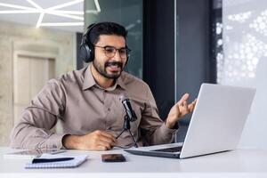 empresario grabación formación en línea curso, hombre sonriente en hablando utilizando profesional micrófono y computadora portátil, trabajando dentro oficina, exitoso negocio entrenador, y financiero. foto