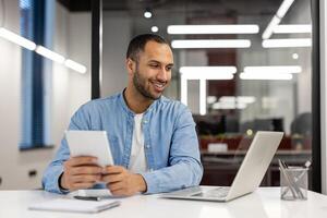 A young Hispanic man is talking on a call on a laptop, sitting at a desk in the office, looking at the screen with a smile and holding a tablet in his hands. photo