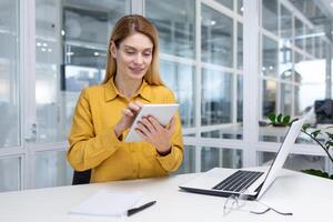 maduro mujer jefe dentro el oficina a el escritorio usos un ordenador portátil y un tableta computadora, un mujer de negocios es satisfecho con el financiero resultados y navega en línea paginas en el Internet. foto