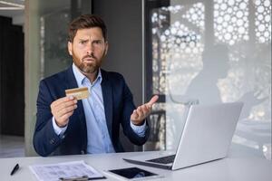 Portrait of a worried and disappointed young businessman man holding a credit card in his hands and seriously looking at the camera while spreading his hands, sitting in the office at the desk. photo
