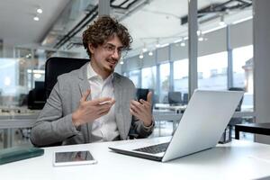 Curly-haired male professional in business attire actively communicating during an online business meeting in a well-lit modern office. photo