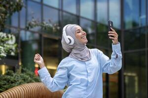 A modern Muslim woman in a hijab is capturing a selfie with her smartphone outside a reflective glass office building. photo