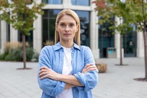 Serious blonde woman in blue shirt standing alone near glass corporate building with hands folded on chest. Successful business owner feeling confident in career development and opportunities. photo