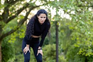 un enfocado Hispano mujer pausar a descanso y captura su aliento durante un intenso rutina de ejercicio en un lozano verde parque a puesta de sol. foto